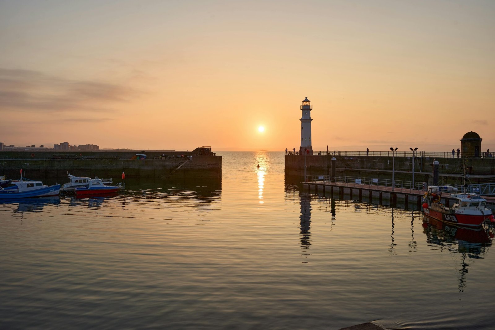 Scenic sunset over a harbor with a lighthouse and boats, reflecting on the calm sea.