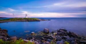 Beautiful view of Cape Auguet Lighthouse during sunset with calm ocean and rocky shore.