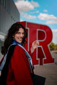 A joyful graduate poses in front of Rutgers University's iconic red R statue.